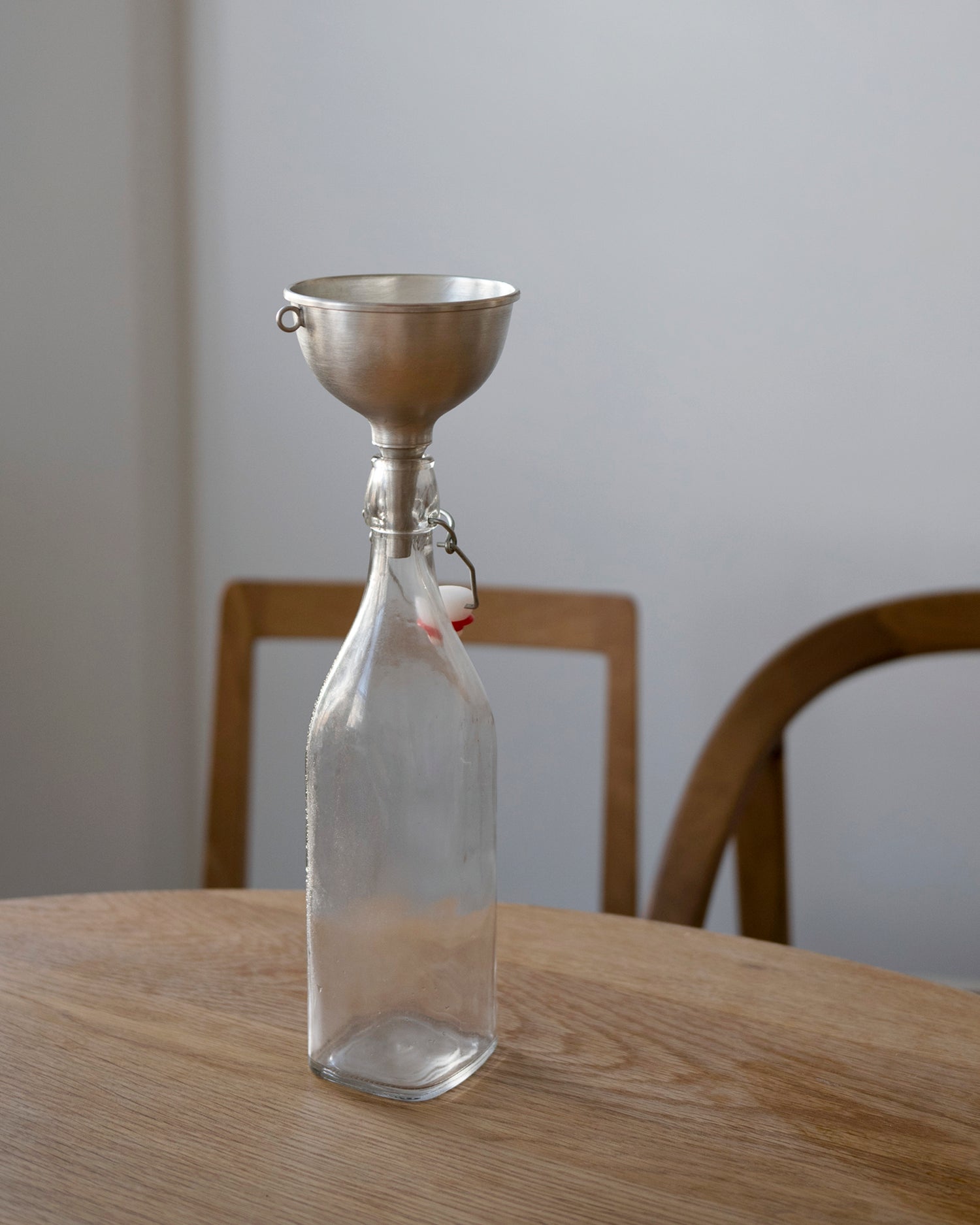 Clear glass bottle with metal funnel by Azmaya on a wooden table against a white wall.