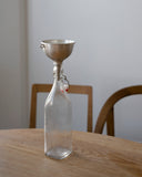 Clear glass bottle with metal funnel by Azmaya on a wooden table against a white wall.