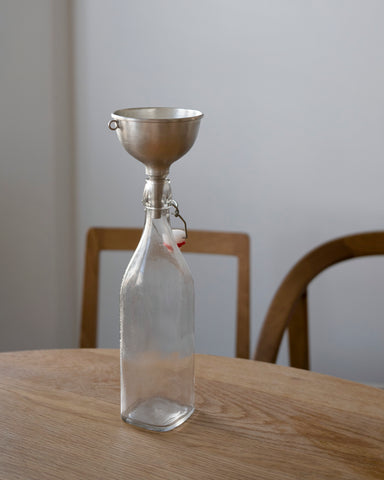 Clear glass bottle with metal funnel by Azmaya on a wooden table against a white wall.