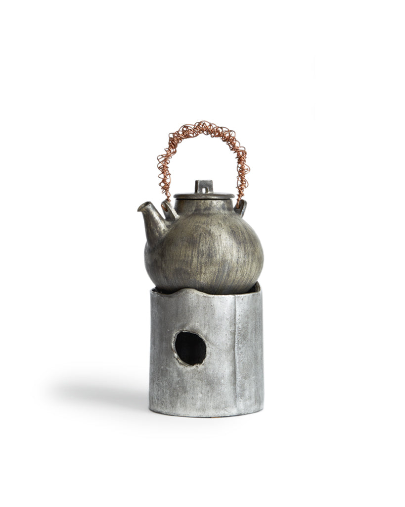 In situation image of ceramic teapot with silver overglaze and twisted copper handle on top of ceramic teapot warmer with sterling silver overglaze by Masanobu Ando against white background.