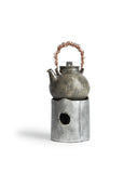 In situation image of ceramic teapot with silver overglaze and twisted copper handle on top of ceramic teapot warmer with sterling silver overglaze by Masanobu Ando against white background.