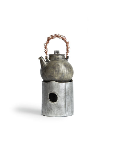 In situation image of ceramic teapot with silver overglaze and twisted copper handle on top of ceramic teapot warmer with sterling silver overglaze by Masanobu Ando against white background.