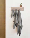 Body, hand, and face zen charcoal towels are hanging on a white oak hanger on a white wall. A wood vase is placed on top of the hanger.
